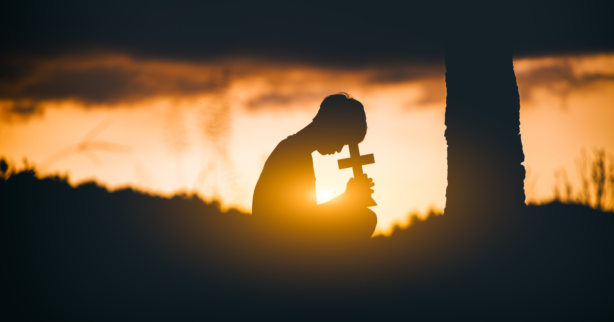 in silhouette, person bows over a small cross