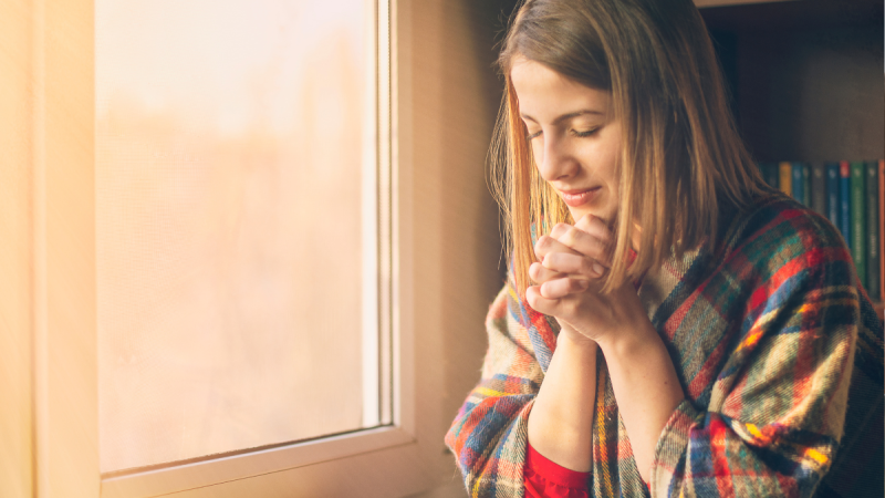 A smiling woman prays next to a sunny window