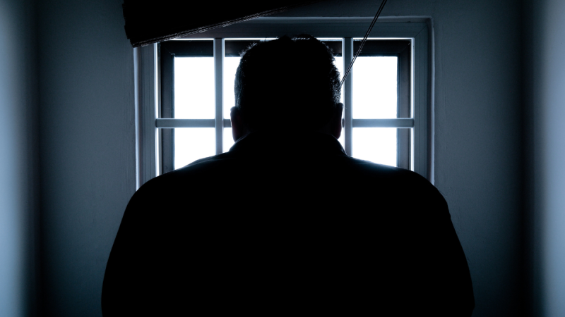A person in a cell stares out through a barred window