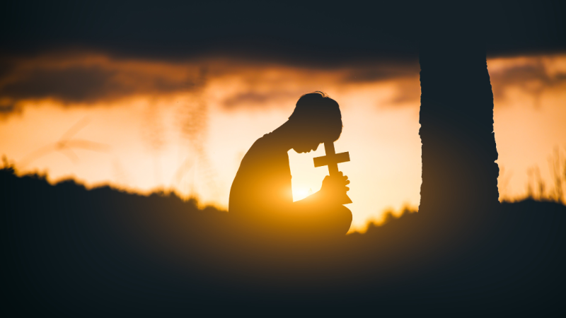 in silhouette, person bows over a small cross
